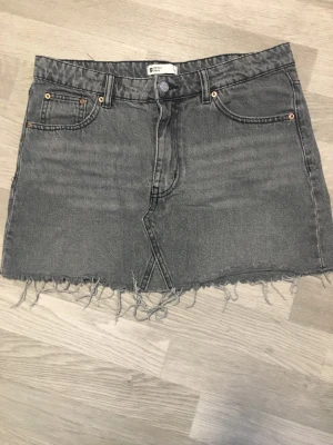 Grå jeanskjol från Gina Tricot - Säljer en grå jeanskjol från Gina Tricot med rå, fransig kant nertill och klassiska fem fickor. Kjolen har bälteshällor och knappstängning framtill. Perfekt för dig som gillar en avslappnad och trendig look.