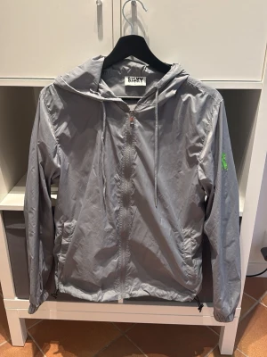 Grå vindjacka från Risky med huva - Säljer en grå vindjacka från Risky i storlek S. Jackan har huva med grönt tryck, dragsko i nederkant och dragkedja framtill. Långärmad modell i lätt polyester, perfekt för blåsiga dagar. Snygg streetstil med enkel design och cool detalj på huvan. Använd max 3 gånger.
