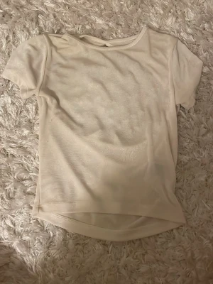 Beige kortärmad topp med öppen rygg - Säljer en beige glittrig topp i mjukt och tunt material. Toppen har korta ärmar och en snygg öppen ryggdetalj med en liten öppning upptill. Passformen är normal och tyget har en lätt glansig finish som ger en fräsch look.
