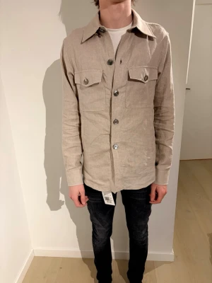 Beige overshirt från Oscar Jacobson - Säljer en stilren beige overshirt från Oscar Jacobson i linne. Storlek S passar M. Köpt för 3000 på Oscar Jacobson.