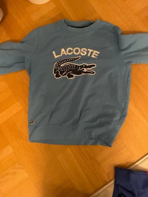 Blå sweatshirt från Lacoste - Säljer en blå sweatshirt från Lacoste med stor krokodillogo och text på bröstet. Tröjan har rund hals, långa ärmar och ribbade muddar. Klassisk och stilren design med broderad krokodil på nederkanten. Perfekt för dig som gillar streetwear.