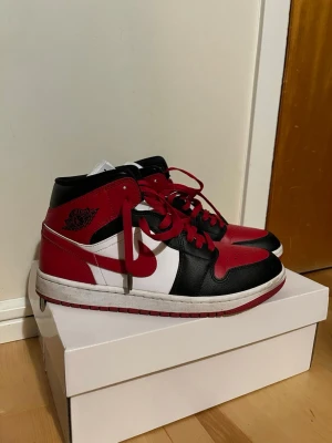 Nike Air Jordan 1 Mid röd/svart/vit - Säljer ett par Nike Air Jordan 1 Mid i klassisk röd, svart och vit färg. Skorna har snygga röda snören, svart och röd ovandel i skinn samt vit sula. Ikonisk Jordan-logga på sidan och hög komfort. Perfekta för dig som vill sticka ut med streetwear-vibe. 