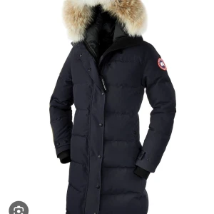Svart dunjacka med päls från Canada Goose - Snygg och varm svart dunjacka från Canada Goose med en lyxig pälsdetalj på huvan. Jackan har långa ärmar och stängs med både dragkedja och knappar. Perfekt för kalla vinterdagar. Har dock ett hål i vänster ficka och vid armhålan men inget som syns 