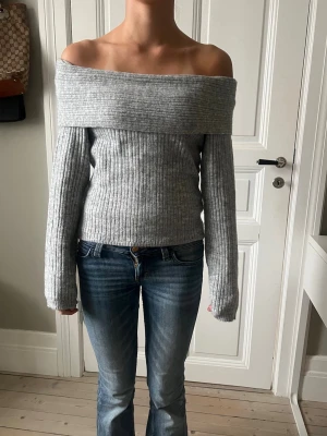 Grå ribbad offshoulder stickad tröja - Säljer en superfin grå stickad tröja med bred offshoulder-ringning. Tröjan är ribbad och har långa ärmar, perfekt för dig som vill ha en trendig och mysig look. Passar till både jeans och kjol för en chill vibe.