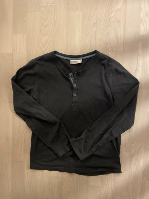 Dexter Morgan Henley :: Dobber - Säljer en mörkgrå långärmad henley från Dobber. Toppen har rund halsringning med tre knappar framtill och en snygg kontrastkant i blått vid knappraden och halsen. Tillverkad i mjuk bomull, perfekt för en avslappnad look, någon som söker archive plagg, eller Dexter fans.   Skavanker: Finns en skavank på sista sliden. Den framstår däremot vara tandkräm eller något liknande, och försvinner med tvätten.  🐕❌ Djurfritt hem 🌿❌ Rökfritt hem  Originalpris: 300 SEK 