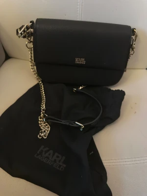 Svart axelväska från Karl Lagerfeld - Snygg svart axelväska från Karl Lagerfeld med guldfärgad kedja och logga framtill. Väskan har en stilren, rektangulär form och är tillverkad i strukturerad syntet med polyesterfoder. Perfekt storlek för det viktigaste och har ett innerfack med mönstrad insida.