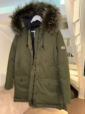 Kenzo grön dunparkas med päls - Säljer en grön Kenzo dunparkas med stor fluffig päls på huvan och svarta detaljer. Jackan har flera fickor, dragkedja och knappar framtill samt Kenzo-logga på ärmen. Insidan är quiltad och fodrad för extra värme. Stilren jacka i fint skick! Använd sparsamt och väl omhändertagen. Endast några små fläckar – därför säljer jag den till ett bra pris. Nypris ca 8 000 SEK – ett superköp för dig som vill ha kvalitet till lägre pris!Storlek L, passar storlek 40-42.Från armhåla till armhåla: 60 cm.L:90 cm