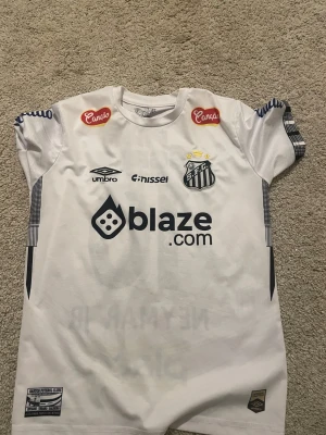 Santos FC Neymar Jr #10 fotbollströja - Snygg vit Santos FC matchtröja från Umbro med Neymar Jr och nummer 10 på ryggen. Tröjan har korta ärmar, tryckta sponsorer och klubbmärke på bröstet. Tillverkad i polyester, med coola detaljer i svart och gult. Perfekt för dig som älskar brasiliansk fotboll och Neymar. Storlek 165-170