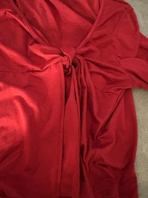 Röd långärmad topp med knyt - Säljer en snygg röd långärmad topp med bred v-ringning och knyt framtill. Modellen är figurnära och har en cool detalj med knuten som ger extra stilpoäng. Materialet känns mjukt och stretchigt, perfekt för dig som gillar att sticka ut med färg.