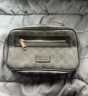 Axelväska  - Snygg grå axelväska från Gucci med klassiskt GG-mönster och svart läderdetalj runt dragkedjan. Väskan har en justerbar axelrem i marinblått och rött, samt en praktisk dragkedja framtill. Perfekt för dig som vill ha en stilren och lyxig accessoar.