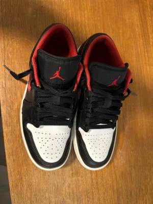 Nike Air Jordan 1 Low svart/röd - Nike Air Jordan 1 Low i svart, rött och vitt med klassisk Jumpman-logga på tungan och Air Jordan-broderi på hälen. Skorna har snörning, perforerad vit tå och röd insida. Swoosh i rött på sidan och platt sula. Tillverkade i skinn och syntetmaterial. Pris kan diskuteras
