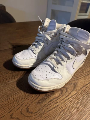 Nike Dunk High vit sneakers - Nike Dunk High sneakers i helvitt och beige med klassisk design och högt skaft. Skorna har snörning, perforerad tå och rund sula. Tillverkade i skinn och syntetmaterial med gummisula. Perfekt för dig som gillar clean och sportig stil.