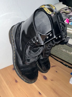 Svarta Dr. Martens boots i skinn - Säljer ett par klassiska svarta Dr. Martens boots med glansig finish och gula sömmar runt sulan. Skorna har snörning, rund tå och platt sula. Tillverkade i slitstarkt skinn och har den ikoniska fliken bak med gul text. Perfekt för dig som gillar edgy stil. Skorna har en del skavanker men är utan hål nya inlägg behövs.