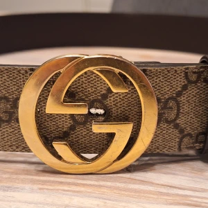 Gucci GG Supreme bälte med guldspänne - Säljer ett Gucci GG Supreme bälte med ikoniskt dubbel-G spänne i guld. Bältet är beige och brunt med det klassiska GG-mönstret och har en bred, rak form. Materialet är skinn med canvas-ytan och detaljer i mörkbrunt. Perfekt för dig som gillar lyxiga accessoarer och vill sticka ut.