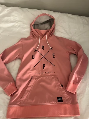 Rosa jackhoodie från Dope  - Säljer en rosa hoodie från Dope med svart grafiskt tryck på bröstet och vit snörning i huvan. Tröjan har en stor magficka, ribbade muddar och är fodrad med mjuk fleece på insidan. Perfekt för skidåkning. Som ny, använd fåtal gånger🤗