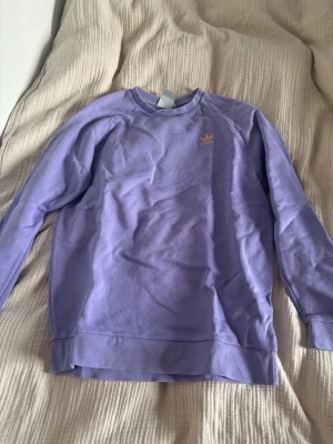 Ljuslila sweatshirt från Adidas - Säljer en ljuslila sweatshirt från Adidas med klassisk logga i gult på bröstet. Tröjan har rund hals, långa ärmar och är gjord i mjuk bomull. Perfekt för dig som gillar sportig och chill stil.
