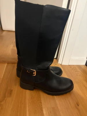 Svarta höga boots med spänne - Snygga svarta boots med högt skaft och guldfärgat spänne vid ankeln. Gjorda i skinn med grov sula och låg klack, perfekt för en edgy look. Klassisk rund tå och dragkedja på insidan för enkel påtagning.