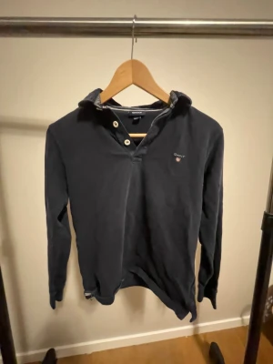 Mörkblå pikétröja från GANT - Snygg mörkblå pikétröja från GANT med klassisk krage och tre vita knappar vid halsen. Tröjan har lång ärm och GANT-logga broderad på bröstet. Perfekt för dig som gillar stilrena och tidlösa plagg.