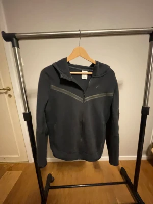 Nike Tech - Svart hoodie från Nike i storlek XL, med huva och halv dragkedja framtill. Snygga detaljer med två diagonala svarta ränder över bröstet och diskret Nike-logga. Mjuk bomullskänsla och långärmad modell, perfekt för chill eller träning.