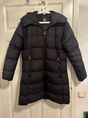 Svart lång pufferjacka från Vinson - Säljer en svart lång pufferjacka från Vinson med huva och tvåvägsdragkedja. Jackan har quiltad design, två fickor med dragkedja och en clean look som passar perfekt när det är kallt ute. Materialet är syntet och jackan är riktigt skön och varm. Den har bara använts 1 gång och är som ny 
