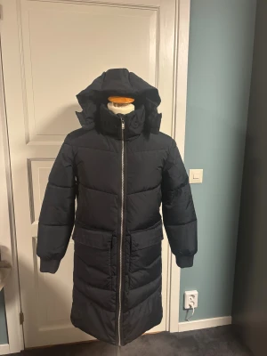 Svart lång pufferjacka med huva - Säljer en svart lång pufferjacka med stor huva och breda kanaler. Jackan har två stora fickor framtill med lock och en rejäl dragkedja som går hela vägen upp i kragen. Perfekt för kalla dagar och har en clean, modern look. Ny skick använt 2 ggr.
