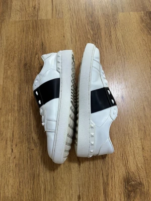 Valentino Garavani vita sneakers - Snygga vita sneakers från Valentino Garavani med bred svart läderdetalj över vristen och coola nitar längs sulan. Skorna har rund tå, platt sula och klassisk snörning. Perfekta för dig som vill ha en lyxig och stilren look med edge.