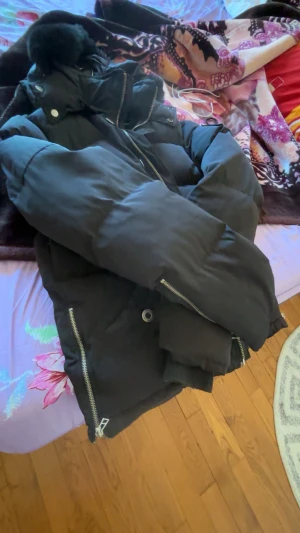 Svart pufferjacka med pälsluva - Säljer en svart pufferjacka från Moose Knuckles med fluffig päls på luvan, flera dragkedjor och knappar. Jackan har lång ärm, ribbade muddar och coola detaljer på fickor och ärmar. Perfekt för dig som vill ha en varm och snygg vinterstil.