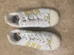 Adidas Superstar Adicolor sneakers - Säljer ett par vita Adidas Superstar Adicolor sneakers med ljusgula ränder och klassisk snäcktå. Skorna har färgglad Adicolor-detalj på sulan och mönstrat innerfoder med pennmotiv. Tjocka vita snören och platt sula. Perfekta för dig som gillar retro vibes och färg.