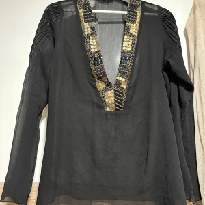 Svart blus med guldpaljetter - Svart blus med lång ärm och djup v-ringning dekorerad med guldiga paljetter och pärlor. Ärmarna har snygga veckade detaljer upptill och tyget är lätt och transparent. Perfekt för dig som vill sticka ut med coola detaljer.