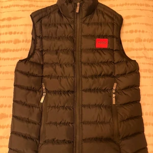 Svart dunväst från Hugo Boss - Svart quiltad dunväst från Hugo Boss med röd logotyp på bröstet och ryggen. Västen har ståkrage, två fickor med dragkedja och en snygg, sportig look. Materialet är polyester och dun, perfekt för lager-på-lager under kyliga dagar.