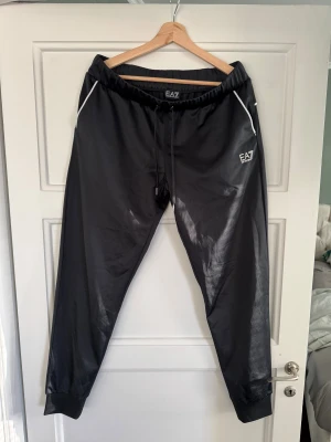 Svarta EA7 mjukisbyxor med snörning - Svarta mjukisbyxor från EA7 Emporio Armani med vita detaljer vid fickorna och bak, samt snörning i midjan. Byxorna har en loose passform och är tillverkade i ett syntetmaterial som ger en lätt glansig look. Perfekta för chill eller träning.