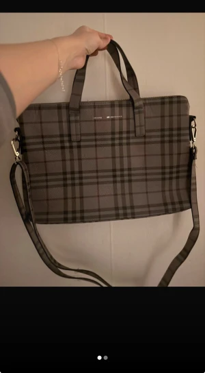 Rutig datorväska från Don Donna - Snygg datorväska från Don Donna i rutig design med grå och bruna toner. Väskan har både handtag och en justerbar axelrem, samt dragkedja upptill. Perfekt för att bära laptop och andra grejer på ett stilrent sätt.