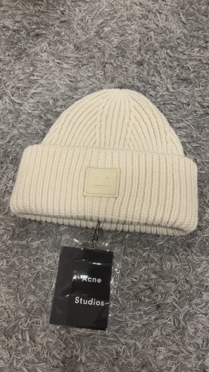 Acne studios mössa - Snygg vit mössa från Acne Studios med ribbad stickning och klassisk logopatch framtill. Mössan är tillverkad i mjuk ull och har en bred uppvikt kant för extra stilpoäng. Perfekt för att hålla dig varm och se trendig ut under kyliga dagar. 