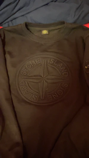 Stone Island Sweatshirt – Svart - Säljer en svart Stone Island-tröja i klassisk design. Tröjan har det stora kompass-emblemet ton-i-ton präglat på bröstet, vilket ger ett stilrent och exklusivt utseende. På vänster ärm sitter det ikoniska Stone Island-märket med knappar, i grönt och gult broderi.