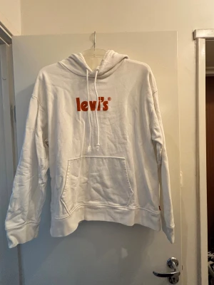 Vit hoodie från Levi's med röd logga - Säljer en vit hoodie från Levi's med röd logotyp på bröstet. Tröjan har huva med snörning och en stor magficka framtill. Mjuk och skön bomullskvalitet, Långärmad och loose passform för en avslappnad stil.