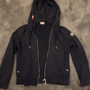 Marinblå Moncler hoodie med dragkedja - Marinblå hoodie från Moncler med huva och snygga snören med röd, vit och svart detalj. Tröjan har lång ärm, Moncler-logga på vänster arm och hel dragkedja framtill. Tillverkad i mjuk bomull för skön känsla och stilren look.