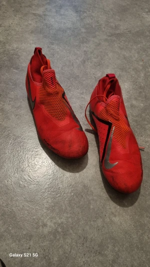 Röda Nike Phantom Vision fotbollsskor - Säljer ett par röda Nike Phantom Vision fotbollsskor med silverfärgade swoosh-loggor på sidorna. Skorna har en strömlinjeformad design med meshpaneler för ventilation och snörning som döljs under ett lager för extra precision. Perfekta för dig som vill sticka ut på planen.