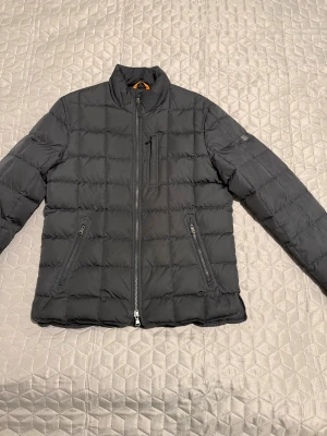 Whitby Lt Down Jacket Morris - Svart pufferjacka från Morris i storlek medium. Jackan har quiltad design, hög krage och tre praktiska fickor med dragkedja. Materialet är syntet och jackan har Morris-logga på ärmen. Perfekt för kyliga dagar och har en stilren look. Jackan är knappt använd och mycket fin. Köpt för 3000kr.