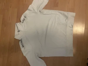 half zip tröja Michael Kors - Vit långärmad tröja från Michael Kors med half zip och hög krage. Snyggt tryck med 'MICHAEL KORS 81' på axlarna. Mjuk insida och klassisk passform, perfekt för en clean och stilren look.