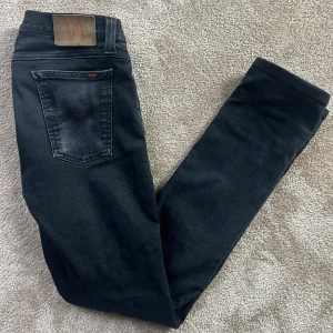 Nudie jeans - Säljer ett par svarta jeans från Nudie Jeans med klassisk design och snygga detaljer på bakfickorna. Storleken på jeansen är W30 L32. Jeansen har en slim passform och är tillverkade i mjukt denimtyg med lätt tvättad utseende. Priset kan diskuteras