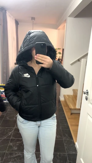 Svart pufferjacka Nike Therma-Fit - Svart pufferjacka från Nike med Therma-Fit-teknologi. Jackan har huva med snörning, dragkedja framtill och vita Nike-loggor på bröstet och insidan. Snygg quiltad design och långärmad modell, perfekt för kalla dagar.