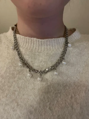 Silverfärgat halsband med pärlor - Snyggt halsband i silverfärgad metall med grov kedja och hängande vita pärlor. Kedjan har en chunky look och pärlorna ger en cool kontrast. Perfekt för dig som gillar att sticka ut med accessoarer och vill ha något som känns både trendigt och klassiskt. Rostfritt stål och handgjort