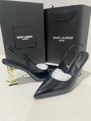 Saint Laurent svarta pumps med YSL-klack - Svarta pumps från Saint Laurent med glansig finish och spetsig tå. Klacken är låg och i guld med ikoniska YSL-loggan som design. Skorna har öppen häl med rem och är tillverkade i skinn. Perfekt statement piece för dig som gillar lyxiga detaljer.