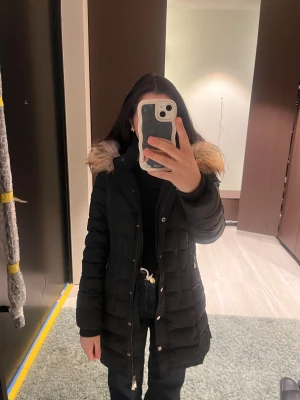 Svart lång dunjacka med päls - Säljer min svarta dunjacka från Svea med fluffig päls på huvan och snyggt emblem på ärmen. Jackan är quiltad, har dragkedja och knappar framtill samt två fickor med dragkedja. Perfekt för kalla dagar och har en stilren, lång passform.