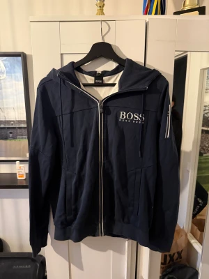 Marinblå hoodie från Hugo Boss - Snygg marinblå hoodie från Hugo Boss med vit dragkedja och logga på bröstet. Tröjan har huva med strukturmönster och praktiska fickor framtill. Perfekt för dig som gillar stilren streetwear med premiumkänsla.