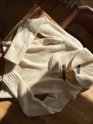 Beige stickad tröja från Polo Ralph Lauren - Snygg beige stickad kofta från Polo Ralph Lauren med bred ribbad krage och muddar. Koftan har en cool knäppning med läderdetalj framtill och långärmade ärmar. Perfekt för dig som gillar klassisk stil med en twist. Stl L i etiketten men sitter mer som en M/38. Inga skador🤍