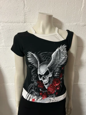 Svart och vit topp med dödskallemotiv - Unik svart och vit topp från Spiral Direct med ett iögonfallande dödskallemotiv med vingar och röda rosor. Toppen har en offshoulder-design och korta ärmar, perfekt för en edgy stil.
