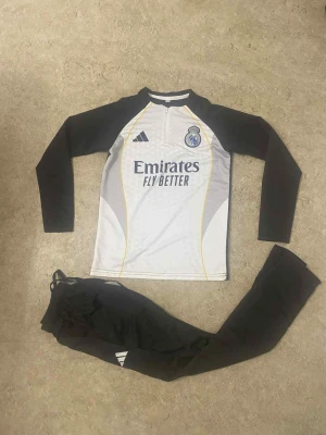 Real Madrid långärmad fotbollströja Adidas - Säljer en långärmad Real Madrid fotbollströja från Adidas i vit och svart med gula detaljer. Tröjan har klubbmärke och sponsortryck på bröstet samt dragkedja vid halsen. Materialet är lätt och andas, perfekt för träning eller match.