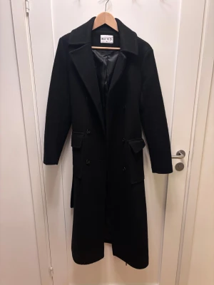 Svart lång kappa från NA-KD - Snygg svart kappa från NA-KD i storlek 38. Lång modell med klassisk krage, dubbla knapprader och två fickor framtill. Bältet med metallhål ger en cool touch och markerar midjan. Perfekt för dig som gillar stilrena och tidlösa plagg.