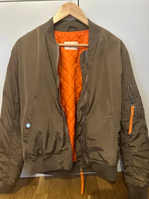 Bomberjacka med orange foder - Står XL men är mer som M i storlek. Snygg bomberjacka med quiltat orange foder och coola detaljer som dragkedja på ärmen med orange band. Jackan har ribbade muddar, silverfärgade dragkedjor och tryckknappar på fickorna. Perfekt för dig som gillar streetstyle och vill sticka ut.